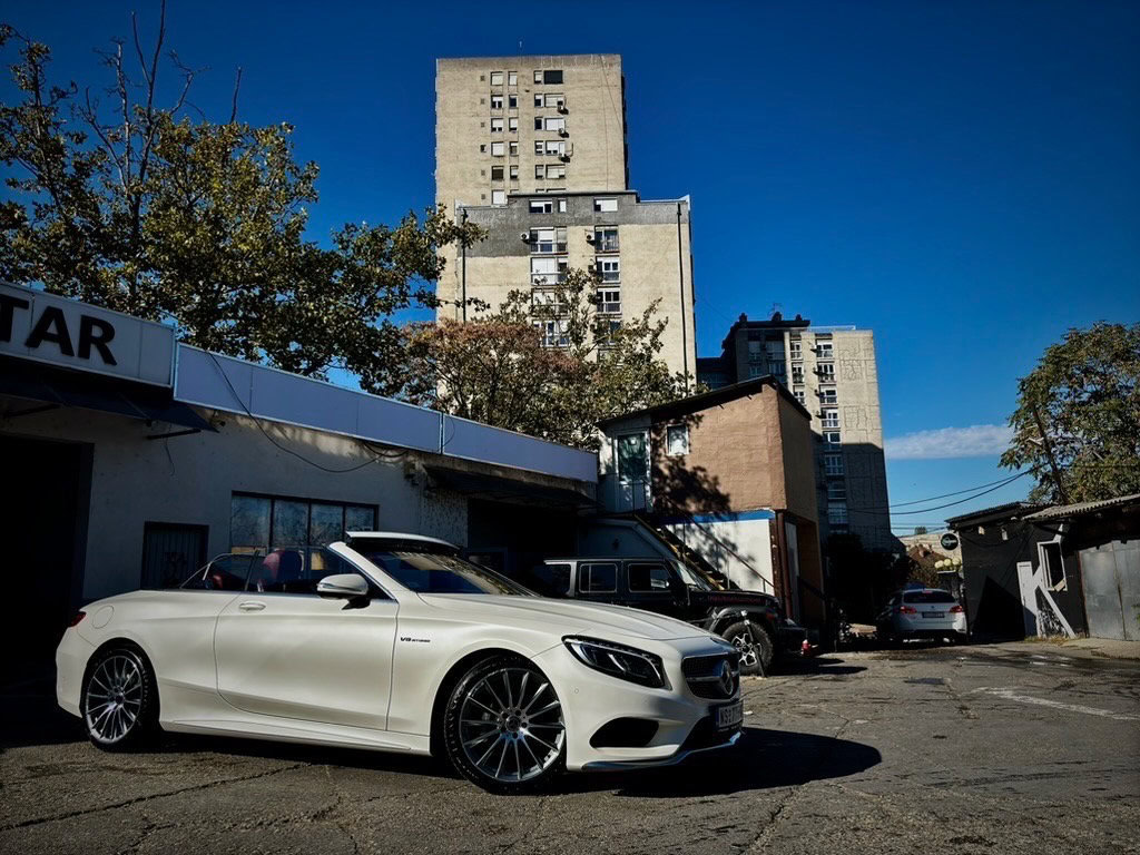 Vozilo Mercedes-Benz kabriolet parkirano pored starog poslovnog objekta u urbanom okruženju, sa visokim višespratnicama u pozadini i plavim nebom.